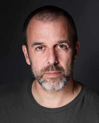 Laurent, cheveux courts et barbe poivre et sel, sur fond sombre.